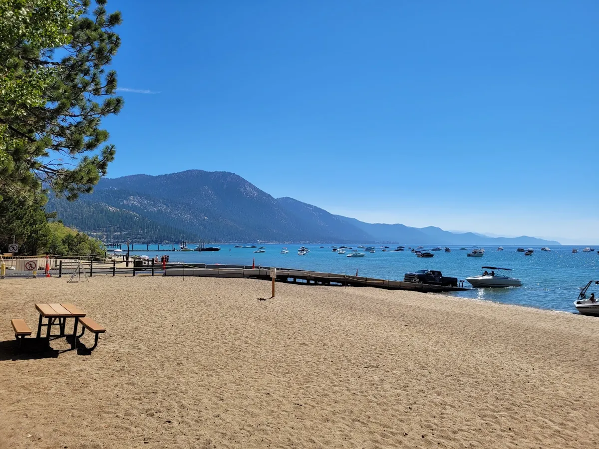 Ski Beach on a clear day: sand, a picnic table, the boat ramp and boats on moorings, the mountains across the lake.