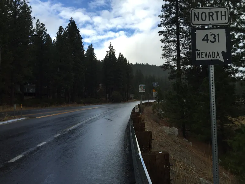 A wet Mt Rose Highway curves past pines under a breaking storm sky, with the North 431 Nevada route sign in the foreground.