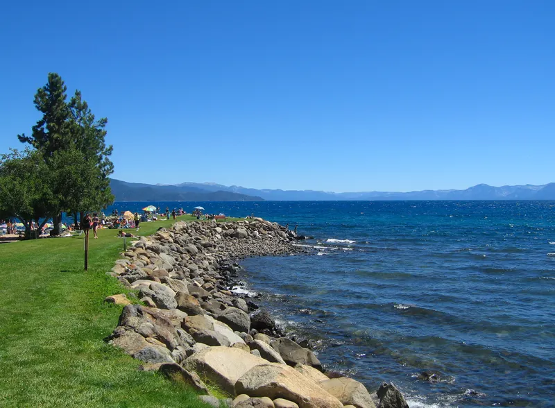 A grassy lawn and a rock wall along the Burnt Cedar Beach shore, with deep blue Lake Tahoe beyond.