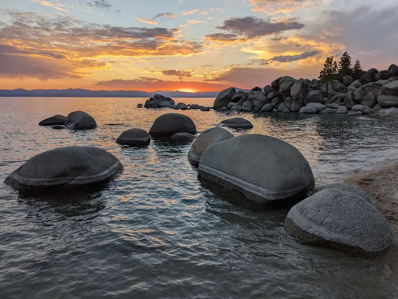 Round boulders in calm water at Chimney Beach under an orange sunset, with rocky points along the east shore of Lake Tahoe.