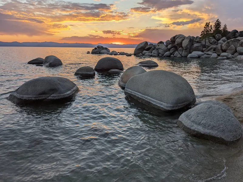 A sandy curve of the Chimney Beach shore at sunset, with big boulders in the shallows and colored clouds over the lake.