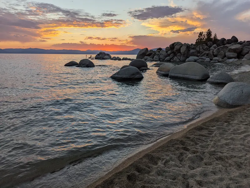 Boulders and a sand beach at Chimney Beach as the sky turns orange and pink over Lake Tahoe.