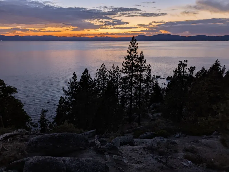 Pine trees frame a dusk view over still Lake Tahoe from the Chimney Beach shore.