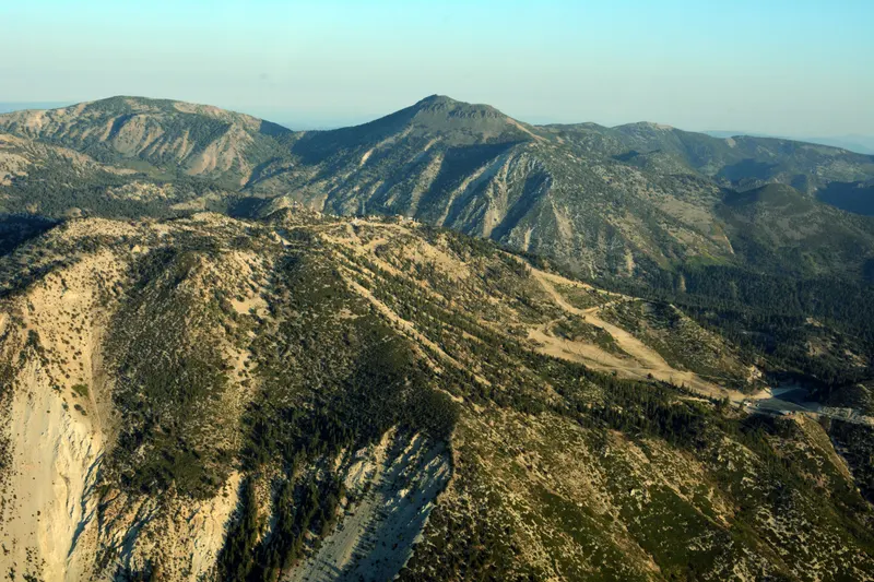Hazy summer aerial of the Diamond Peak ski area, with bare ski runs cut through dry, forested mountains and no snow.