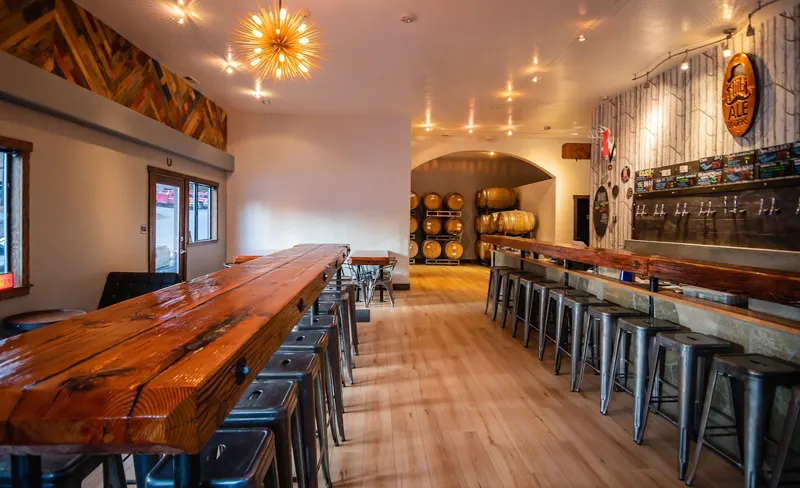A long wooden bar with metal stools and a wall of barrels behind it.