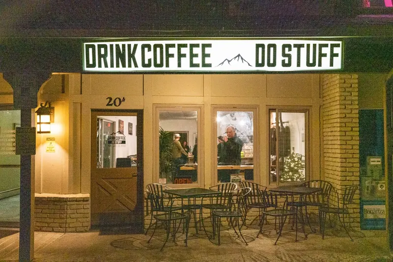 The Drink Coffee Do Stuff storefront lit at dusk, with patio tables out front.
