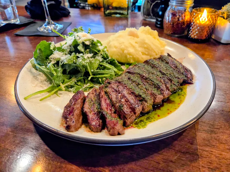 Sliced grilled steak with green herb sauce, mashed potatoes and an arugula salad with shaved cheese, on a wood table by a candle.