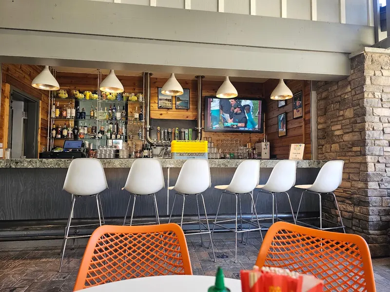 A bar with white stools and orange chairs under pendant lights.