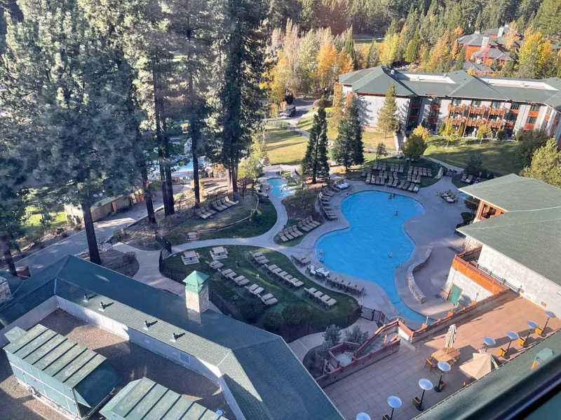 An aerial view of a lakeside resort pool, lawns and low buildings among tall pines in fall color.