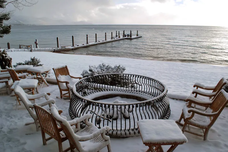 A snowy lakeside deck with chairs around a fire ring and a pier over the water at Lake Tahoe.
