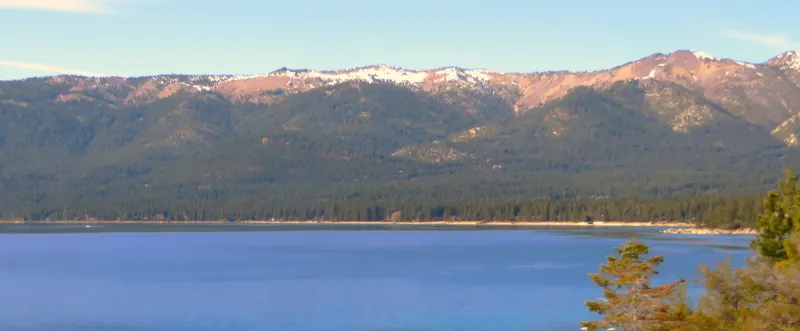 Deep blue Lake Tahoe below forested mountains with snow on the peaks, seen from the Incline Village shore.
