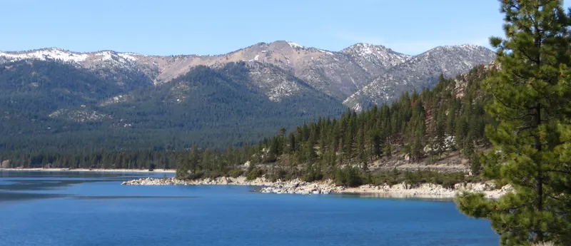 Deep blue Lake Tahoe with a forested point and snow-dusted peaks across the water.