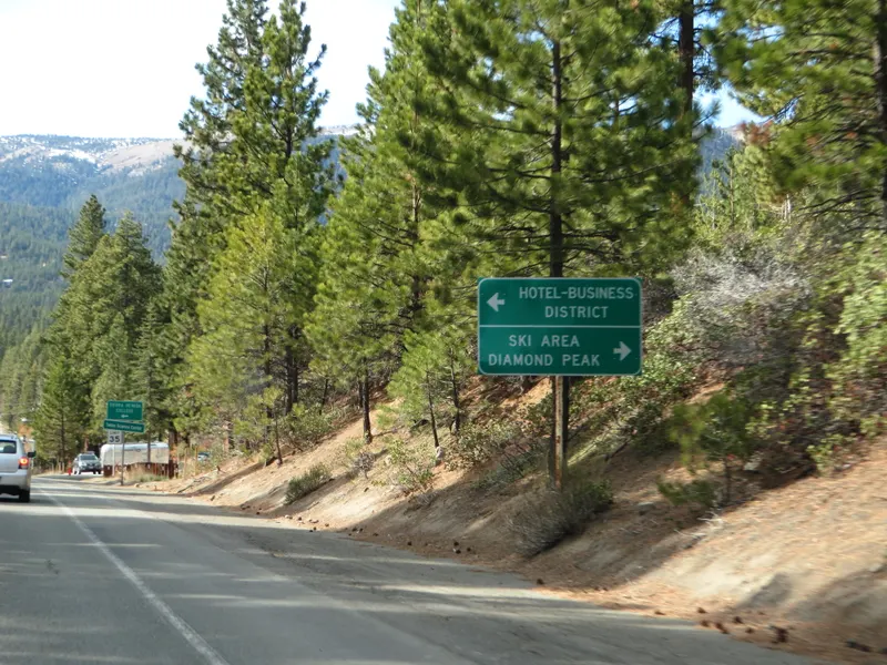 Nevada State Route 28 with a green sign pointing to the Hotel-Business District and the Diamond Peak ski area, among pines.