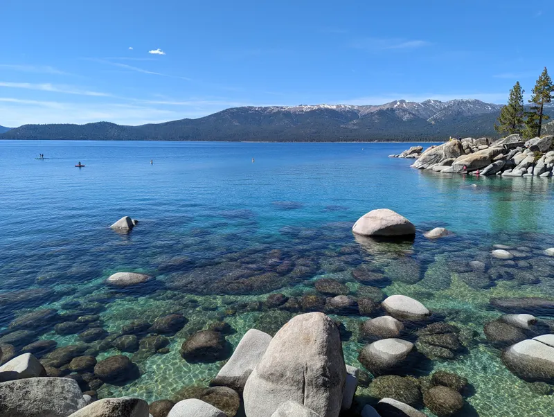 Clear water over pale boulders at Sand Harbor, with paddleboarders on the lake and a rocky point beyond.