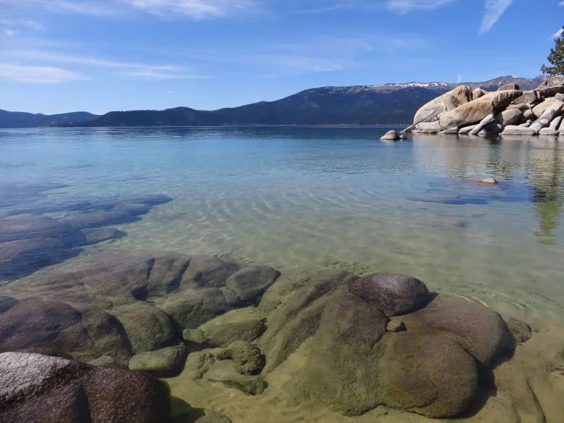 Clear turquoise shallows over rounded boulders at Sand Harbor, with a granite island and snowy mountains beyond.