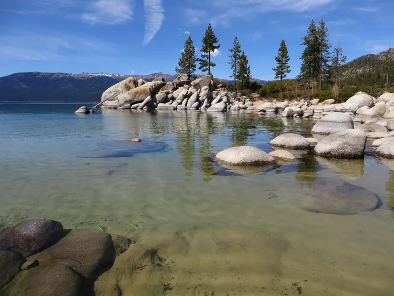 A granite island and pines reflected in clear, shallow water at Sand Harbor, with snow on the mountains across the lake.